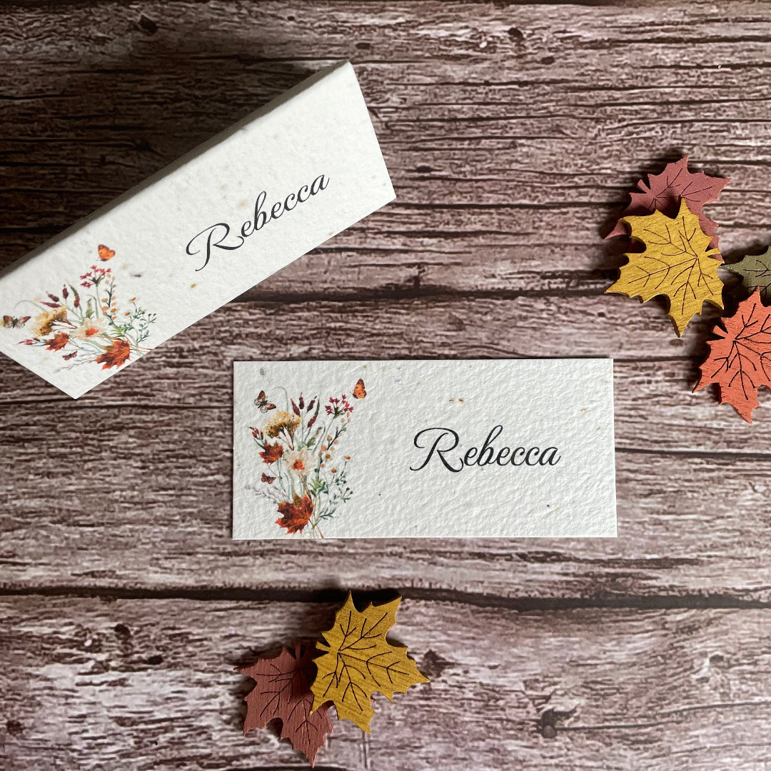 Personalized name card with floral design on a wooden surface with decorative autumn leaves and wildflowers.