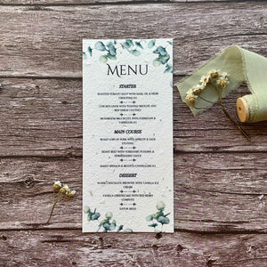 A wedding menu card with a eucalyptus theme, featuring plantable seed paper and printed text, displayed on a wooden surface with a ribbon and decorative eucalyptus leaves.