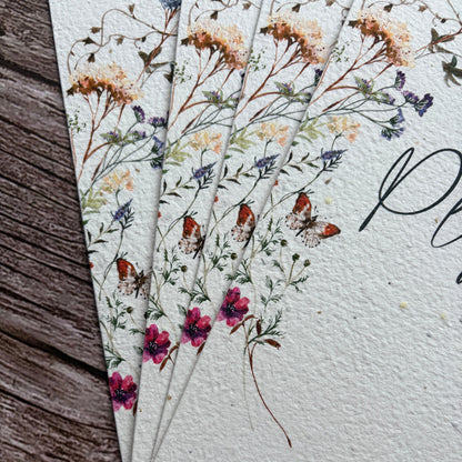 A close-up of several plantable table names cards with delicate watercolor floral and butterfly illustrations, arranged on a rustic wooden surface.