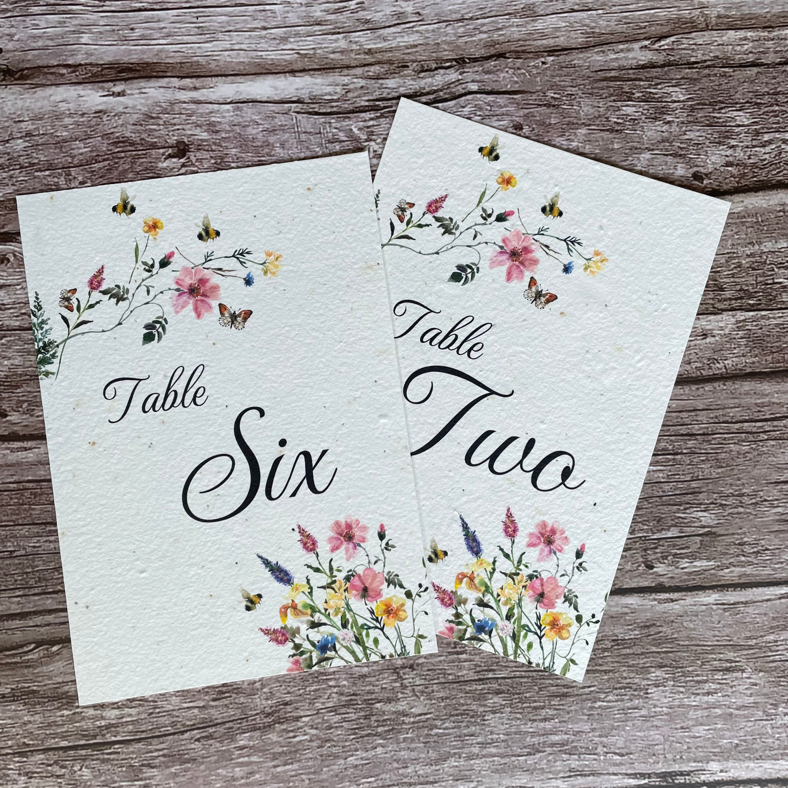 Two floral table number cards on a wooden surface made of seed paper