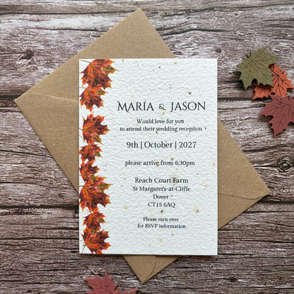 A plantable wedding invitation card with a vertical border of orange and red autumn leaves, placed on top of a brown kraft envelope on a rustic wood background with scattered fall leaves.