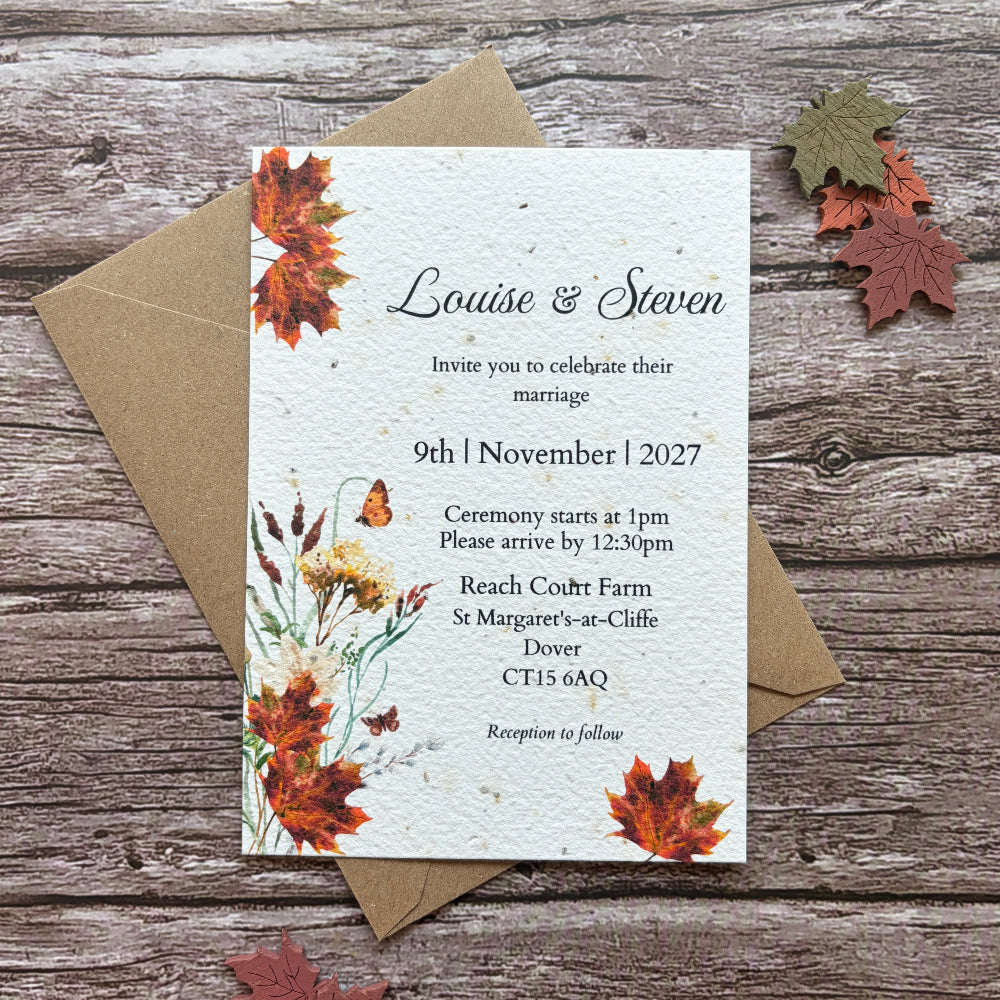 A plantable autumn wedding invitation card with autumn floral illustrations and the names Louise &amp; Steven, placed on top of a kraft envelope on a rustic wood surface with scattered fall leaves around it.