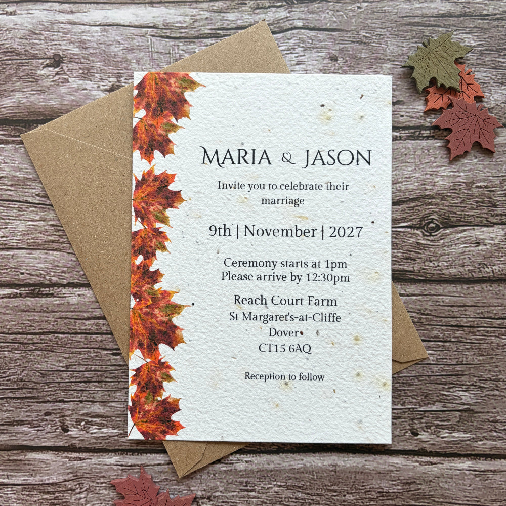 Plantable autumn wedding invitation card with a vertical border of orange and red autumn leaves on the left, placed on top of a kraft paper envelope on a rustic wood surface. Small decorative leaves are scattered around it.