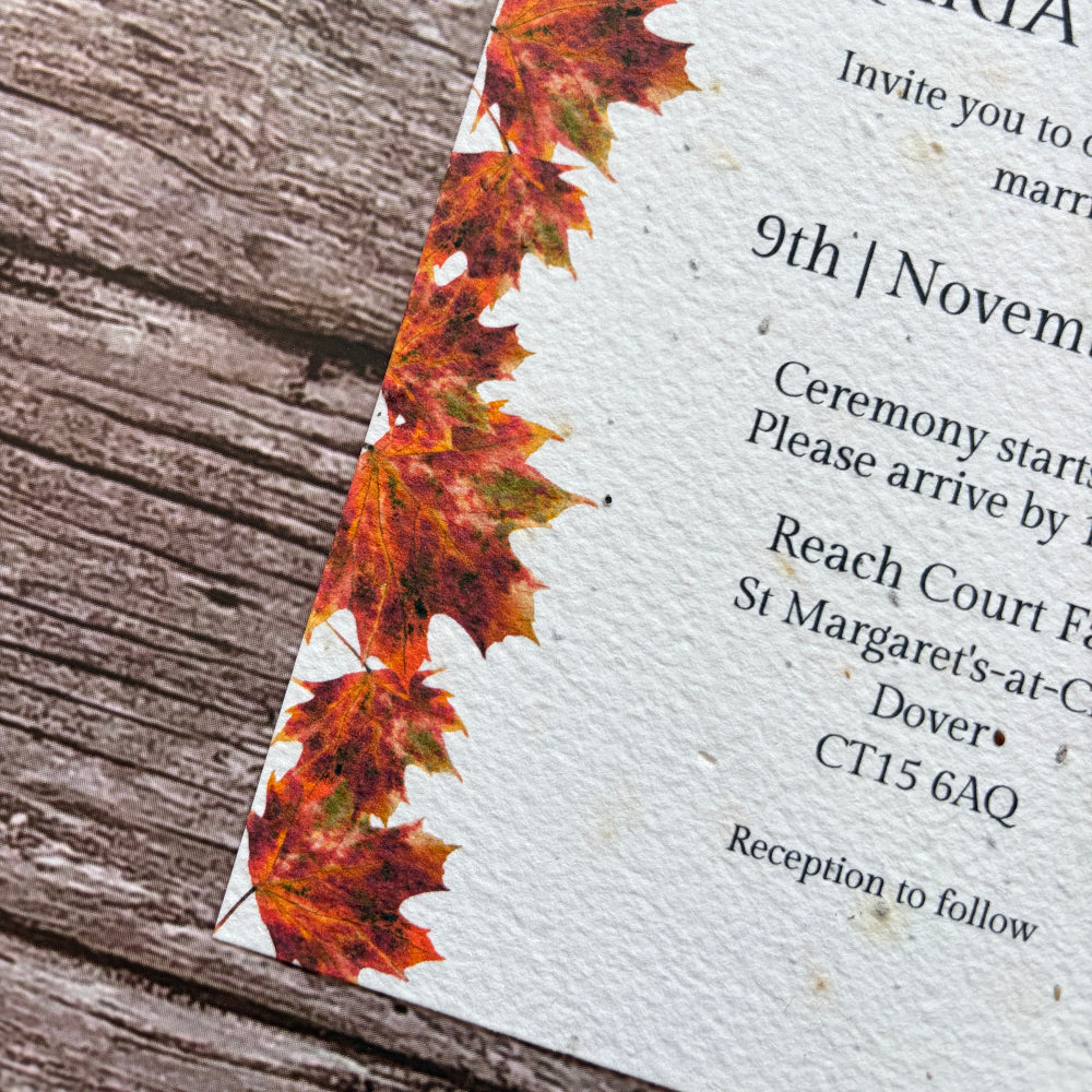 A close-up of a plantable wedding invitation card with a border of autumn maple leaves in orange, red, and brown on a textured white background, placed on a rustic wooden surface.