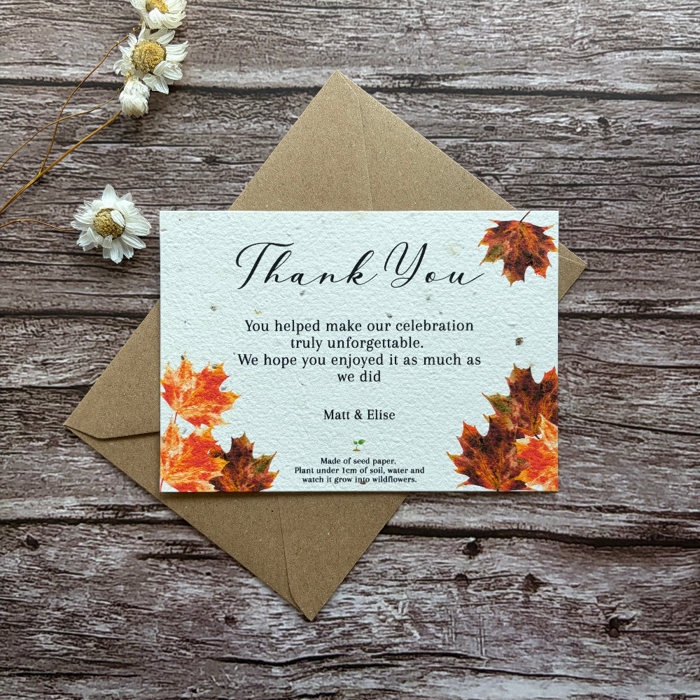 Plantable wedding Thank you card with autumn leaves design on a wooden surface with a brown envelope and white flowers.