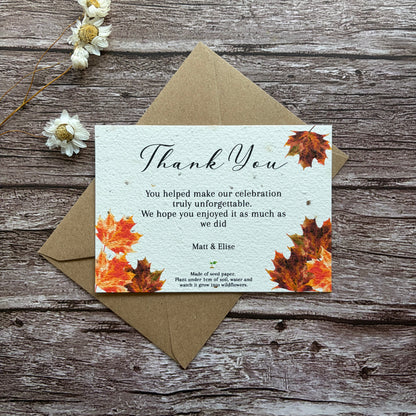 Plantable wedding Thank you card with autumn leaves design on a wooden surface with a brown envelope and white flowers.