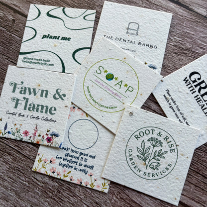 Square business card seed paper samples with various designs and branding on a wooden surface