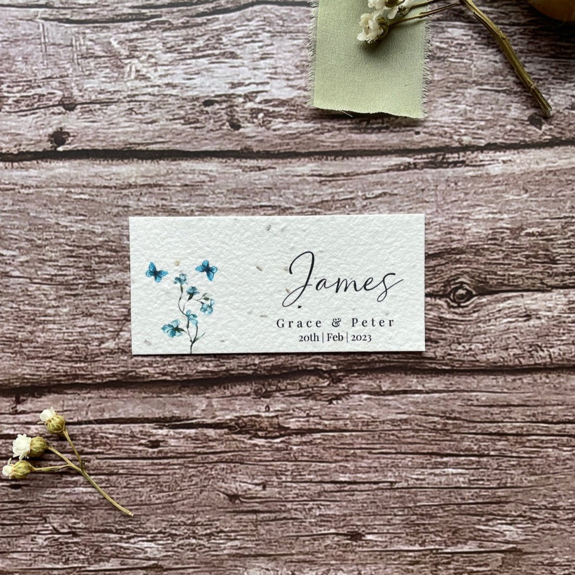 plantable seed paper wedding guest place cards in dusty blue theme, showing a close-up front view, floral design on wooden background, eco-friendly