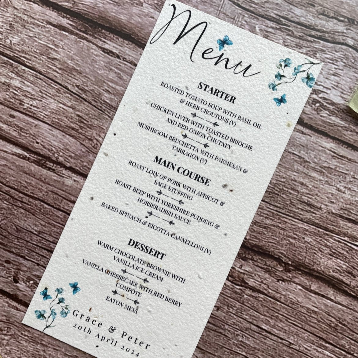 plantable seed paper wedding menu card in dusty blue theme, showing an angled view, floral design on wooden background, eco-friendly