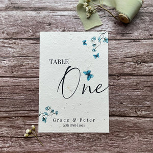 plantable seed paper wedding table number/name card in dusty blue theme, showing front view, floral design on wooden background, eco-friendly.