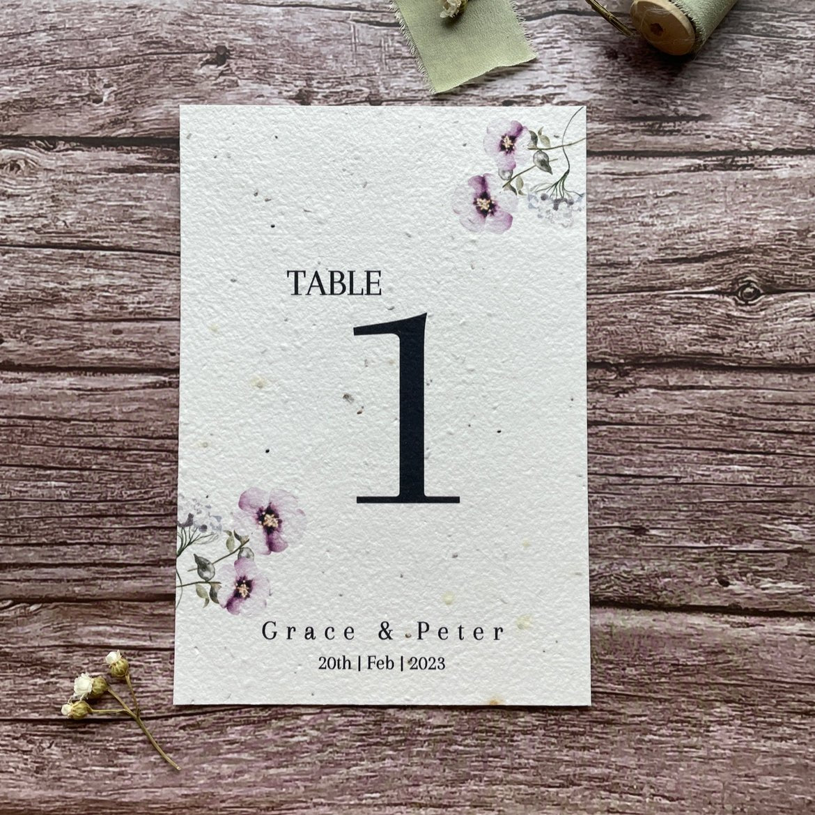 plantable seed paper wedding table number/name card in lilac theme, showing front close-up view, floral design on wooden background, eco-friendly